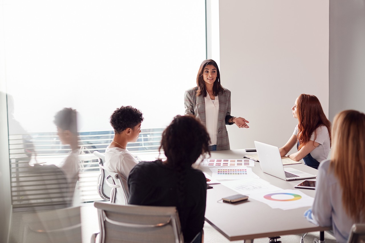 Female Boss Gives Presentation To Team Of Young Businesswomen Meeting Around Table In Modern Office