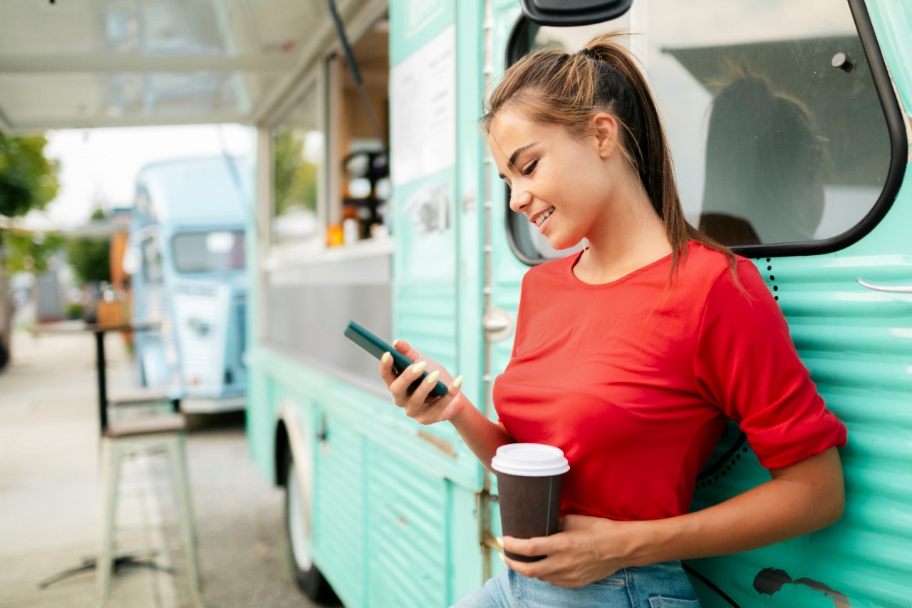 Happy young woman wearing a red shirt, leaning onto an old food truck, having a cup of coffee and using a smart phone, smiling